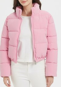 Doudoune rose avec un col haut, un design matelassé et un ourlet élastique. Elle possède une fermeture éclair à l'avant et est portée sur un haut blanc.