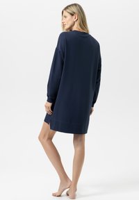 Vrouw die blootsvoets staat, met de rug naar de camera, in een langemouwen, marineblauw sweatshirtjurk met zijsplitten op een witte achtergrond.