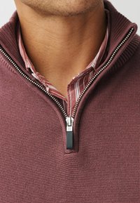 Bordeauxroter Reißverschluss-Pullover aus strukturiertem Stoff mit geripptem Kragen und silbernem Reißverschluss-Detail. Darunter ist ein leicht gestreiftes Hemd teilweise sichtbar.