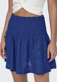 Falda azul de encaje con cintura fruncida, que presenta un dobladillo acampanado y patrones de ojales. Hecha de tela ligera y texturizada.