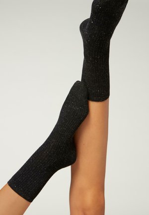 Svarta ribbade strumpor med metalliska prickar, mjuk textur och längd till mitten av vaden, placerade på ben med kontrasterande hudton.