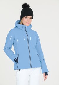Blå skijakke med høj krave, frontlynlås og sidelommer. Har hvide detaljer og en tætsiddende pasform. Sort strikket hat med pompon.