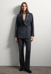 Completo su misura grigio scuro con giacca doppiopetto, pantaloni dritti e scarpe nere a suola piatta. Tessuto liscio con un design strutturato.