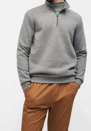 Man draagt een grijze trui met kwart-ritssluiting over een groene coltrui, met handen in de zakken van bruine broek, tegen een effen achtergrond.