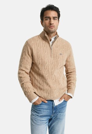 Beige Kabelstrickpullover mit hohem Kragen und Viertelreißverschluss. Gerippte Bündchen und Saum, getragen über einem weißen Hemd mit Kragen. Klassische Passform.