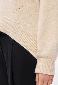 Svetlo béžový pletený sveter s rebrovaným lemom a manžetami, s malými dierkami pre textúru. Logo vyšívané blízko spodného okraja.