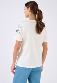 Mujer con cabello castaño en una coleta, vistiendo una camisa blanca de manga corta con un patrón de hojas azules y rosas en la manga y pantalones azules, de espaldas.