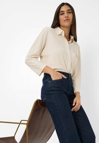 Femme aux longs cheveux bruns portant une chemise beige à col et un jean bleu foncé, se tenant avec une main dans la poche à côté d'un fauteuil en cuir marron.