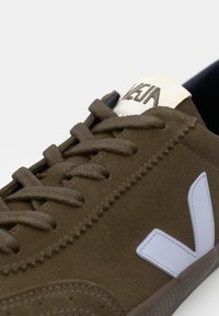 Sapatilha em camurça castanha com o logo V em branco, biqueira redonda, atacadores tonais e forro interior preto, apresentando sola de borracha texturizada e detalhes costurados.