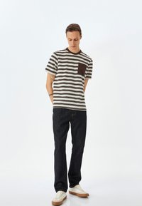 Stribet t-shirt i beige og mørkebrun med en brystlomme, parret med mørke jeans og lyse sneakers. Simpelt, afslappet design.