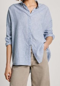 Light blue and white striped button-up shirt with a relaxed fit, rolled-up sleeves, and a collar, paired with beige pants.