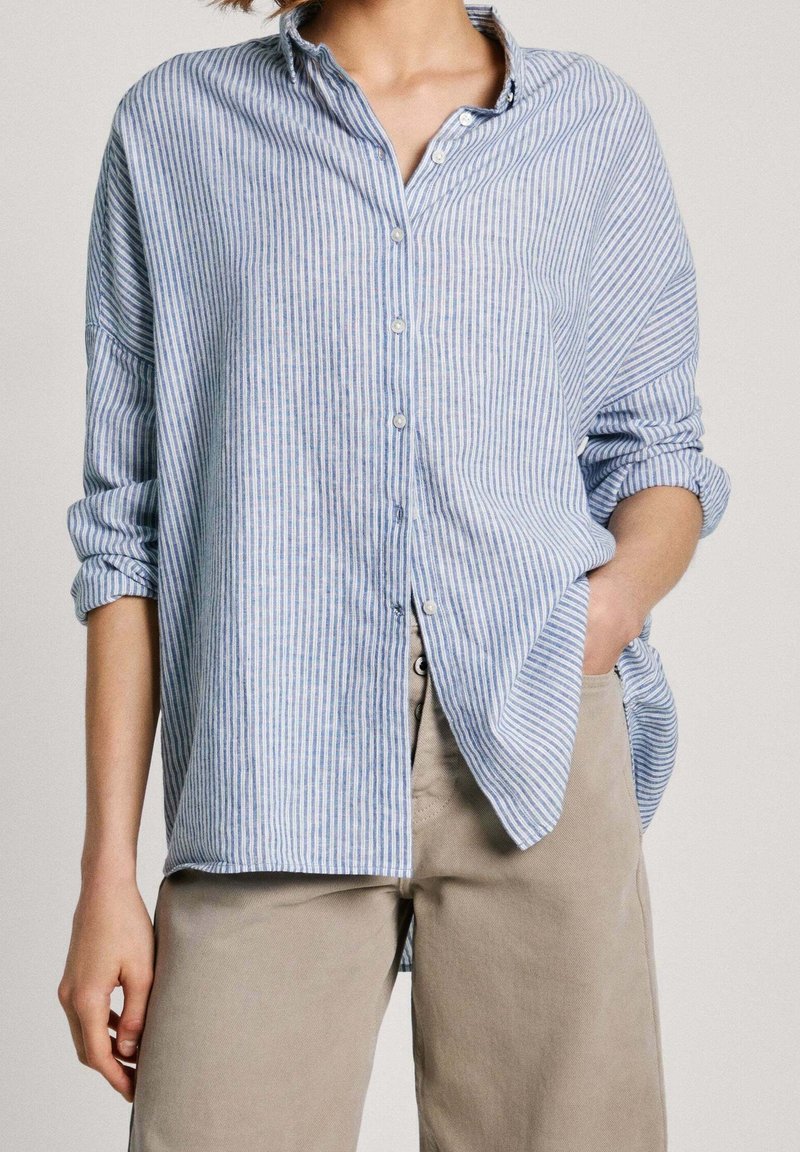 Light blue and white striped button-up shirt with a relaxed fit, rolled-up sleeves, and a collar, paired with beige pants.