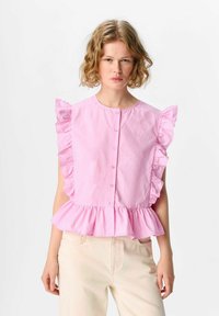 Blusa rosa a balze con maniche corte, scollatura rotonda e chiusura con bottoni. Presenta un orlo a peplo e una texture liscia. Abbinata a pantaloni chiari.