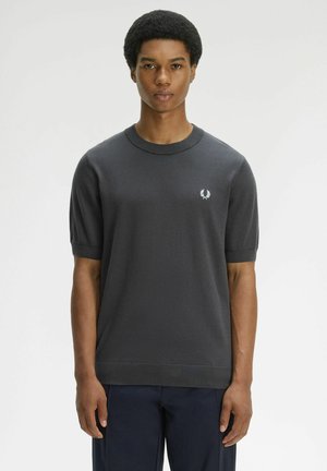 Giovane uomo con capelli corti e ricci che indossa una maglia a maniche corte in maglia grigio scuro e pantaloni blu navy, in piedi davanti a uno sfondo semplice.