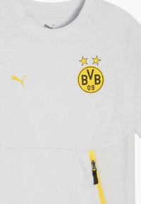 Valge sportlik särk, millel on kollane Puma logo, BVB embleem kollase ja musta värviga, kaks tähte logo kohal ning külgedel kollased tõmblukud.