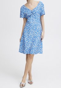 Modré šaty s krátkým rukávem, květinovým potiskem, výstřihem do V a nabíranými detaily na přední straně. Nosené s stříbrnými sandály na podpatku.