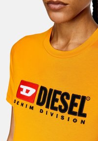 Pomarańczowa bawełniana koszulka z czarnym haftowanym logo, na którym widnieje napis "DIESEL" oraz pod nim fraza "DENIM DIVISION" w czarnym tekście.