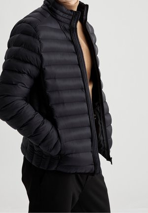 Person trägt eine schwarze gesteppten Daunenjacke über einem beigen Rollkragenpullover, die Hände in den Jackentaschen, vor einem schlichten hellen Hintergrund.