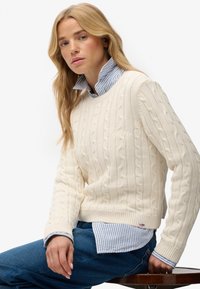 Crème gebreide kabeltrui met geribbelde zoom, laag over een blauw-wit gestreept shirt. Model zit op een houten stoel.