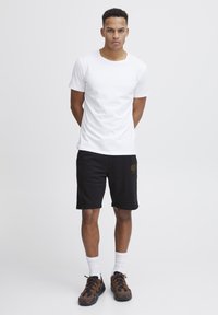 Weißes Baumwoll-T-Shirt kombiniert mit schwarzen Shorts, die ein goldenes Herz-Logo zeigen. Das Modell trägt braune und schwarze Sneaker mit weißen Socken.