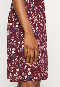 Vínově červené květinové šaty se zářivými růžovými, modrými a bílými květy. Měkká textura s uvolněným střihem. Viditelná ruka, která spočívá na látce.