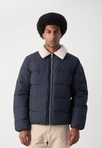 Marineblå pufferjakke med en shearlingkrave, prydet med quiltede detaljer, frontlynlås og afslappet pasform. Blød tekstur, let materiale.