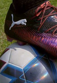 Crampon de football Puma noir et orange, tige texturée, lacets rouges, reposant sur un ballon de football noir et blanc à motifs avec des accents bleus.