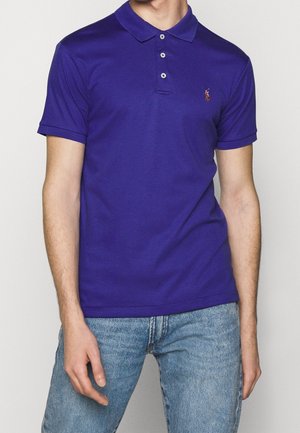 Homme portant un polo violet ajusté à trois boutons avec un petit logo brodé, associé à un jean bleu clair.