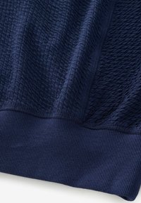 Marineblauwe trui met een geweven stof met verhoogde patronen, ribbelmanchetten en een gladde zoom. Het materiaal lijkt lichtgewicht en ademend te zijn.