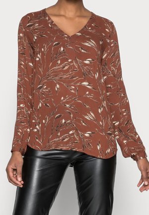 Bluse mit langen Ärmeln in Rostfarbe mit beigem und braunem Blatt-Blumenmuster, getragen zu schwarzen Kunstlederhosen, gezeigt vom Hals bis zur Hüfte.