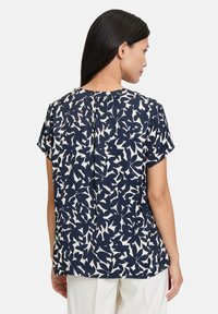 Blusa blu navy con motivo di foglie bianche. Maniche corte, scollatura arrotondata e dettagli con pieghe sul retro. Tessuto leggero e liscio.