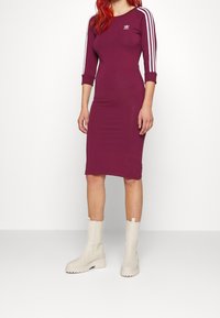 Burgundy klänning med figurnära passform och tre vita ränder längs ärmarna samt en liten logotyp på bröstet. Matchas med gräddfärgade ankelstövlar.