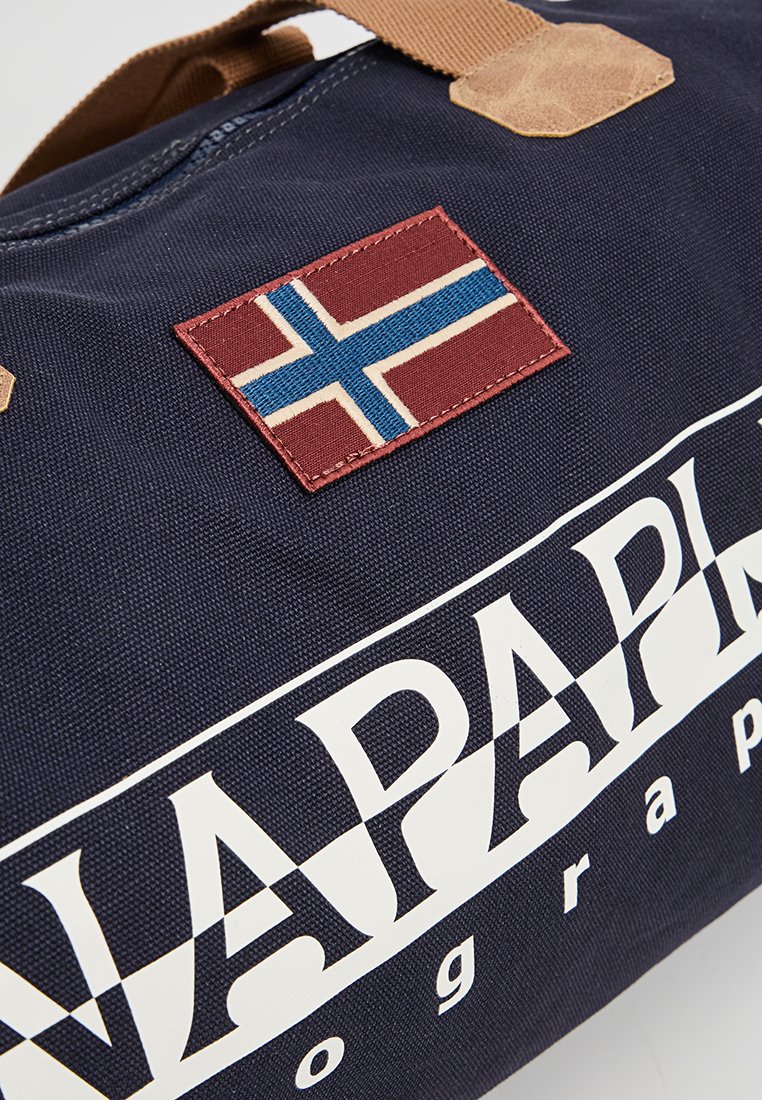 Granatowa torba z brązowym uchwytem, haftowaną naszywką w kształcie nordyckiego krzyża oraz dużym białym, wyraźnym napisem "NAPAPIJRI" z boku.