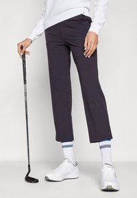 Calças de golfe em azul marinho escuro, feitas de tecido leve, com corte reto. Usadas com ténis brancos e meias listradas a azul.