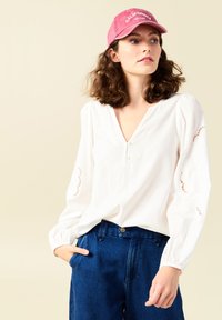 Witte blouse met V-hals en decoratieve kantaccenten op de mouwen, gecombineerd met donkerblauwe denimbroek met hoge taille en een roze pet.
