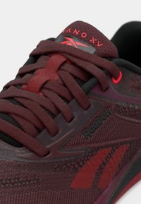 Mörkröd sportskor med texturerad ovandel, framträdande snören och Reebok-logotyp. Dämpning och flexibel sula synlig, med en smidig design.