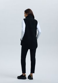 Schwarze Fleece-Weste mit hohem Kragen, lockerer Passform und Seitentaschen, kombiniert mit schwarzen Hosen und Schuhen. Glatte Textur.