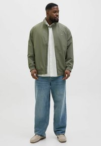 Olive green bomber jacket with a stand collar, worn over a white striped shirt, paired with light blue baggy jeans and beige slip-on shoes.