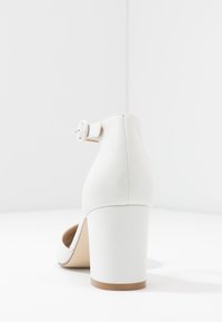 Weißer Absatzschuh mit runder Spitze, Knöchelriemchen mit Schnalle und strukturiertem Lederoberteil. Der Blockabsatz ist robust und tanfarben.