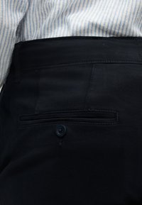 Κοντινό πλάνο μαύρου παντελονιού με κουμπωμένη πίσω τσέπη, φορεμένο με μπλε και άσπρο ριγέ πουκάμισο που είναι μπατζωμένο μέσα.