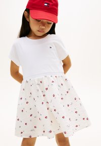 Λευκή κοντομάνικ η μπλούζα με φούστα σε ανοιχτό ύφασμα με λουλουδένιο σχέδιο, διακοσμημένη με κόκκινα και μπλε λουλούδια. Κόκκινο καπελάκι με λογότυπο μπροστά.