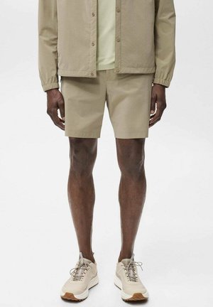 Homme portant un short beige décontracté, une veste assortie et des baskets beiges, debout devant un fond uni.