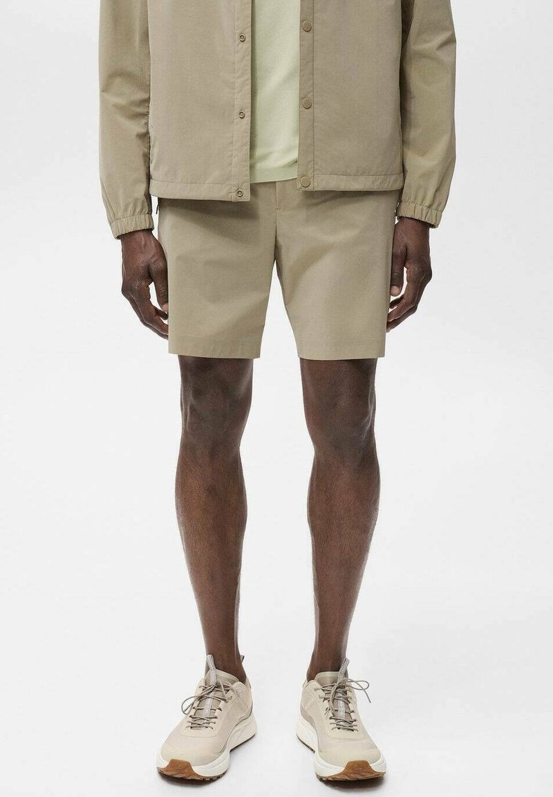 Homme portant un short beige décontracté, une veste assortie et des baskets beiges, debout devant un fond uni.