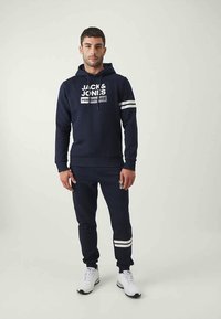 Muž oblečený v tmavomodrom mikinovom svetri Jack & Jones a zodpovedajúcich joggeroch s bielymi pruhmi, stojaci na jednofarebnom svetlom pozadí.