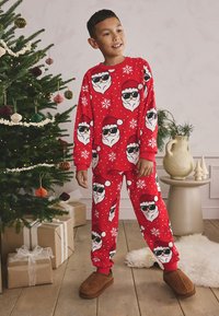Ensemble de pyjama rouge avec des visages de Père Noël portant des lunettes de soleil et des flocons de neige sur un tissu doux. Coupe ample avec manches longues et pantalon assorti.