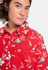 Camisa de botones roja con un patrón de flores y plumas en blanco, gris y negro. Incluye un bolsillo frontal con una etiqueta de logo. Material de algodón.