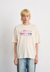 T-shirt en coton beige avec un logo "New Balance" multicolore en rose, rouge et bleu. Manches courtes et coupe décontractée.