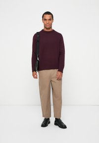 Μπορντό πλεκτό πουλόβερ, κρεμ μπεζ παντελόνι και μαύρα παπούτσια. Το μοντέλο κρατάει μια μαύρη τσάντα χιαστί. Απλή, ίσια σιλουέτα με μίνιμαλ λεπτομέρειες.