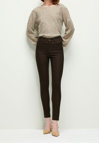 Femme portant un pull beige à manches longues avec des sequins, un pantalon moulant marron foncé et des chaussures slingback beiges à talons, debout contre un mur blanc.