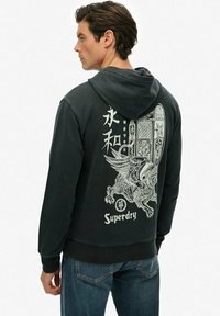 Hombre vistiendo sudadera negra con gráfico blanco de un león alado, caracteres asiáticos verticales y texto "Superdry" en la parte posterior, combinado con jeans azules.